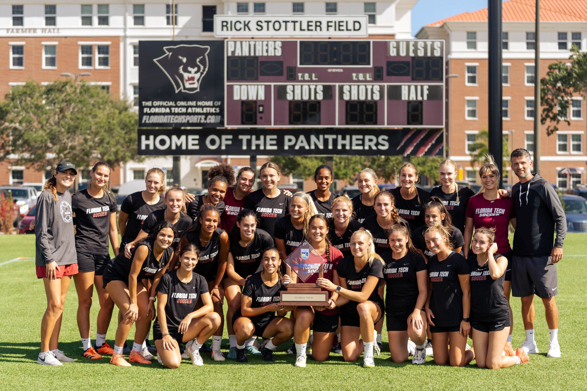 Florida Tech women's soccer team posing together on the field, holding a trophy, with the scoreboard and campus buildings in the background